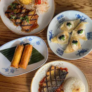 Four plates with grilled fish, rice, spring rolls, sliced grilled meat, salad, and fancy topped dumplings on a wooden table.