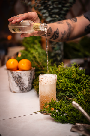 A tattooed arm pours a drink into a frothy cocktail glass, surrounded by greenery and oranges.
