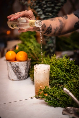 A tattooed arm pours a sparkling drink into a cocktail glass on a counter with greenery and oranges.