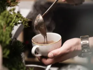 A person pours hot coffee into a white cup, with steam rising and greenery in the foreground.