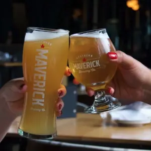 Two people clink glasses of craft beer labeled 'Maverick' in a bar setting.