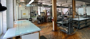 Spacious printmaking studio with large presses, wooden floors, tables, and industrial lighting.