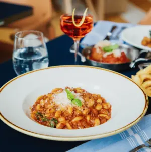 A white plate of pasta with red sauce and cheese, with drinks and food on a table in the background.