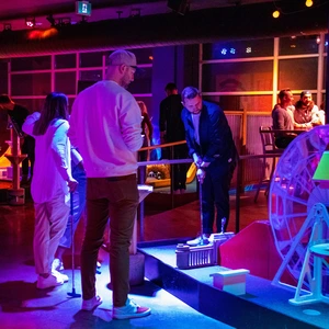 People playing mini golf indoors under colorful, neon lighting with a Ferris wheel decoration nearby.