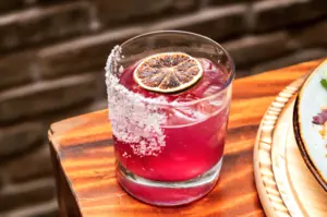 A pink cocktail with ice, a dried lime slice, and a salted rim, served in a glass on a wooden table.