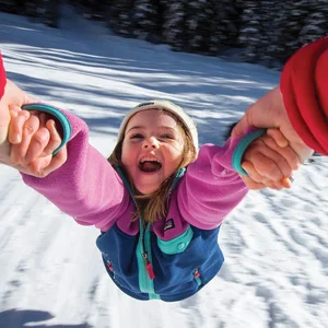 Smiling child in winter clothes being spun around while holding hands with an adult on snowy ground.