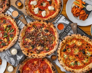 Several wood-fired pizzas with various toppings on a table, with dipping sauces and chicken wings on the side.
