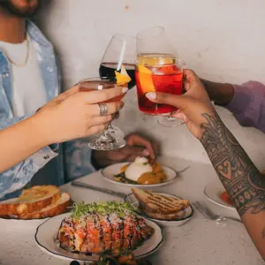 Four people clinking drinks over a table with food, including toast and a dish topped with greens.