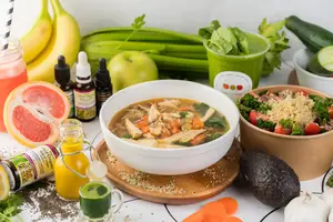 A bowl of soup, salad, fruits, veggies, juice shots, and supplements arranged on a table.