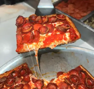 A slice of square pepperoni pizza is being lifted from a pan, with cheese stretching.