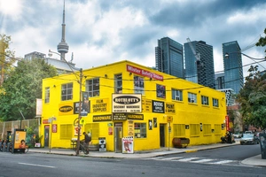 A bright yellow hardware store building on a city corner, with modern skyscrapers in the background.
