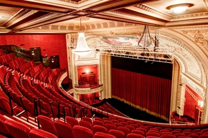 Ornate theater interior with red seats, a chandelier, and a stage with a closed red curtain.
