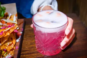 A hand holds a pink cocktail with a frothy top next to a plate of nachos on a wooden table.
