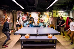 Five people play table tennis in a colorful, graffiti-decorated room with bright lighting and snacks nearby.