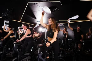 A group of people energetically cycling in a dark studio, waving white towels in the air.