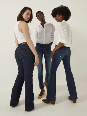 Three women wearing jeans and tops stand posing together against a plain, light-colored background.