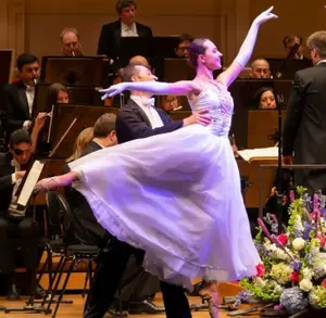 Ballet dancer in a white dress performs on stage with an orchestra and conductor in the background.