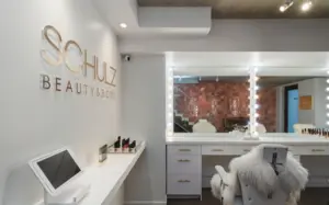 A modern beauty salon with a vanity mirror, white chair, and 'Schulz Beauty Room' on the wall.