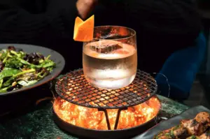 A cocktail with an orange peel garnish sits on a grill above glowing coals, beside a plate of salad.
