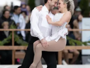 Two ice skaters perform together, smiling, as the woman lifts her leg while held by her partner.