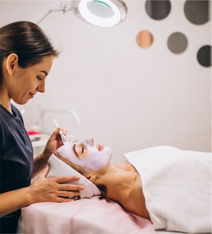 Esthetician applies a facial mask to a client lying down under a lamp in a spa treatment room.