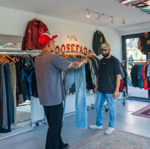 Two men look at jeans inside a clothing store with racks of clothes and jackets on display.