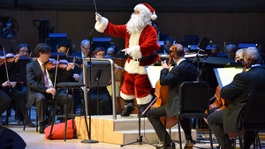 Santa Claus conducts an orchestra on stage, surrounded by musicians playing string instruments.