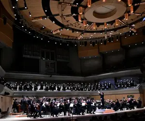 A symphony orchestra and choir perform on stage in a large, modern concert hall.