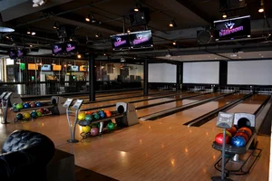 Modern bowling alley with empty lanes, colorful bowling balls, and overhead screens in a dimly lit setting.