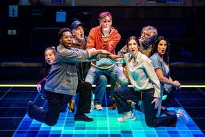 Seven actors onstage, dramatically posing and singing, with bright blue lighting and a grid pattern on the floor.
