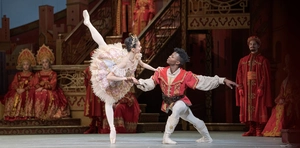 Two ballet dancers perform on stage in ornate costumes, with elaborately dressed performers in the background.