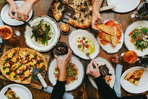 A group of people share pizza, salad, and drinks around a wooden table at a meal gathering.