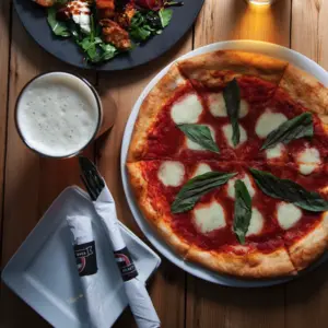 Pizza with basil and mozzarella, a glass of beer, and a salad on a wooden table with utensils nearby.