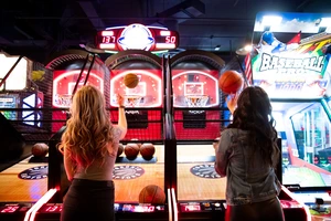 Two women play an arcade basketball game, tossing balls toward the hoops under bright lights.