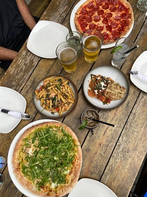 A wooden table with pizzas, pasta dishes, beers, and empty plates set for a meal.
