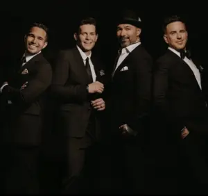 Four men dressed in black suits standing together and smiling against a dark background.
