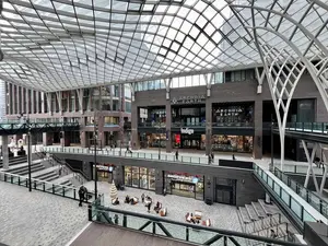 Modern glass-roofed shopping mall with multiple levels, shops, and people sitting on benches in the atrium.