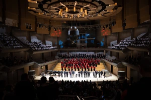 A large choir and orchestra perform on a brightly lit stage in a modern concert hall filled with an audience.