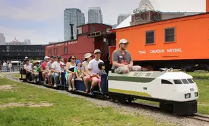 People riding a miniature train outdoors, with city buildings and colorful train cars in the background.