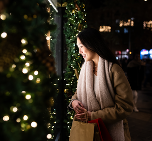 Woman with shopping bags smiles while looking at a holiday window display decorated with lights and greenery.