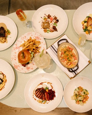 A table set with various plated gourmet dishes, a burger with fries, and several drinks.