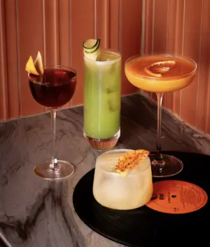 Four colorful cocktails on a marble table, one served on a vinyl record, with garnishes on each drink.