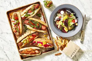 A tray of gourmet hot dogs, a salad bowl, fries in a paper bag, and a small cup of pickles on a marble table.