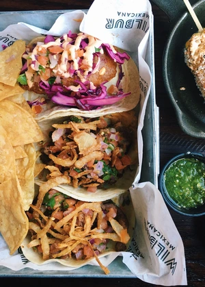 Three tacos with various toppings, tortilla chips, green salsa, and Mexican street corn on the side.