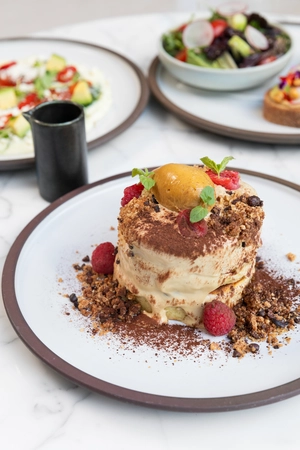 Tall dessert with cream, raspberries, crumble, and mint on a white plate; salad and tart in the background.