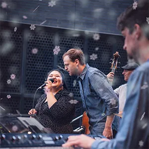 Four musicians perform on stage; one sings into a microphone while others play keyboard, bass, and guitar.