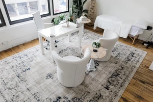 Modern office with white chairs, desk, potted plants, and a patterned rug on wooden floor near large windows.